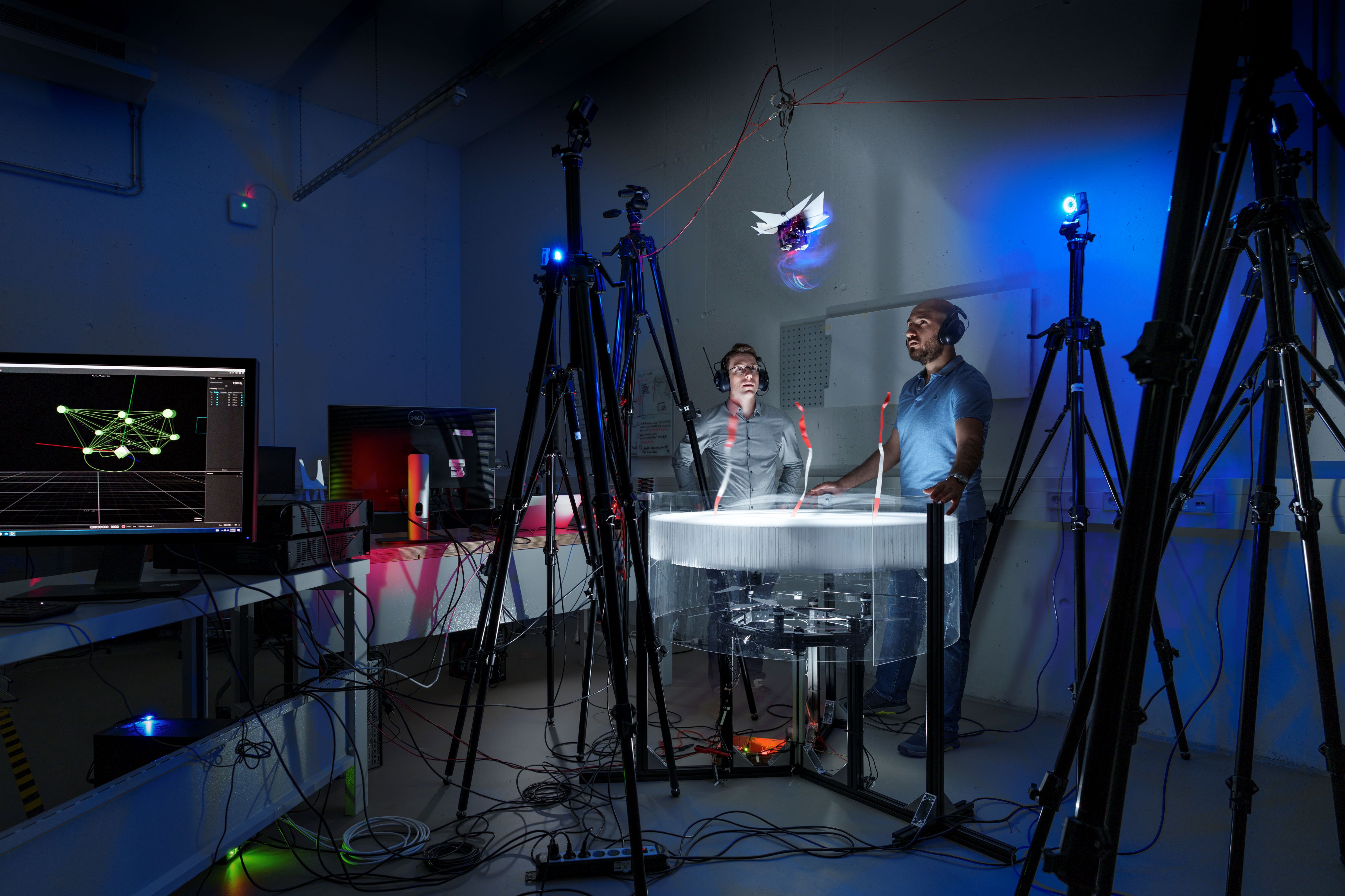 Two people wearing headphones observe a drone hovering above a circular, transparent containment unit filled with a white, swirling substance in a dimly lit laboratory setting with multiple tripods an